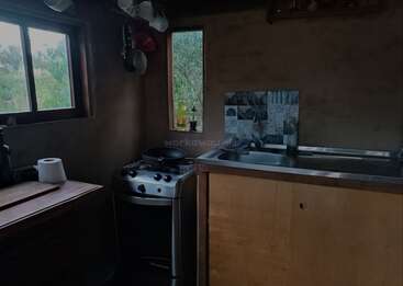 This cozy kitchen features a gas stove, a metal sink, wooden cabinets, cutting board, hanging utensils, small windows, and natural light illuminating the rustic interior space.