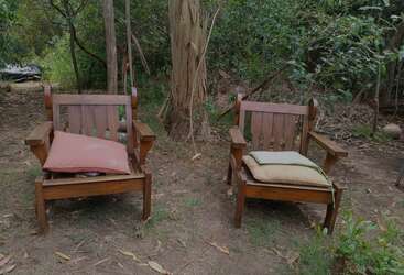Two wooden chairs with cushions are placed outdoors on grass and dirt, surrounded by trees and plants, creating a peaceful and rustic garden setting.