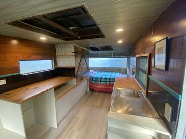 Cette image montre un intérieur de camping-car confortable et rénové avec des panneaux en bois. Il comprend un coin cuisine, un plan de travail, un lit avec une couverture à carreaux, de grandes fenêtres et un éclairage abondant.