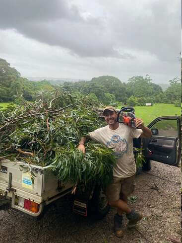 Un homme se tient à côté d'un camion utilitaire chargé de branches, souriant et tenant une tronçonneuse. La scène se déroule en extérieur, avec une végétation luxuriante et un ciel nuageux.