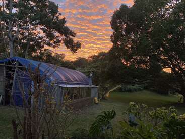 Un bâtiment rustique au toit bleu se dresse au milieu d'une verdure luxuriante et de grands arbres, éclairé par un ciel de coucher de soleil vibrant, rayé d'orange et parsemé de nuages.