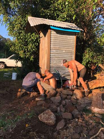 Trois hommes travaillent ensemble, torse nu, construisant ou réparant quelque chose avec des pierres devant un petit hangar en tôle ondulée, entouré d'arbres et de soleil.