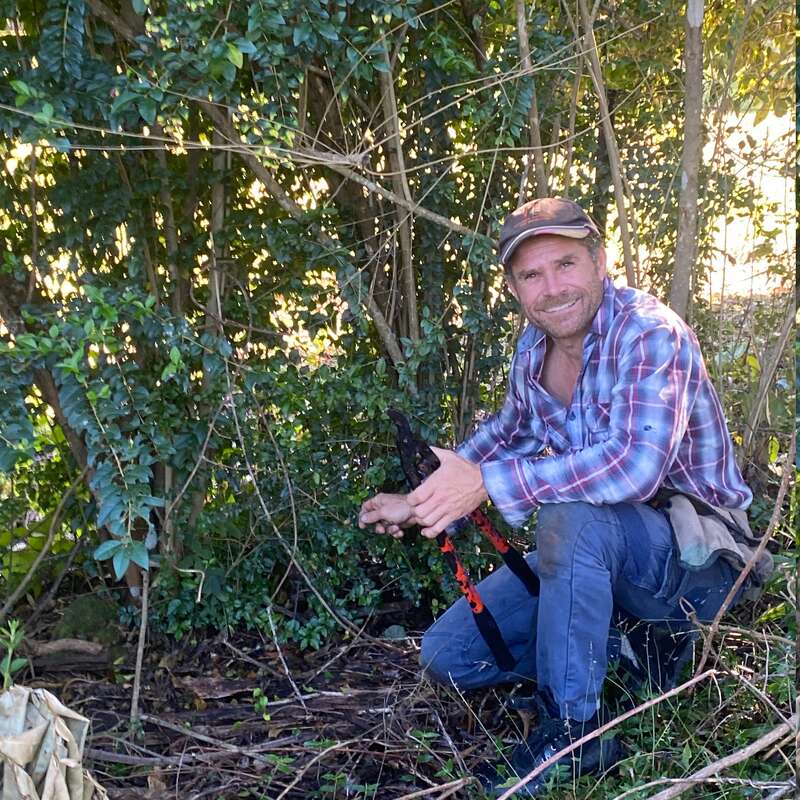 Un homme vêtu d'une chemise à carreaux et d'une casquette est agenouillé à l'extérieur, souriant tout en tenant un sécateur de jardinage. Il travaille au milieu d'un feuillage vert dense, entouré de végétation naturelle.