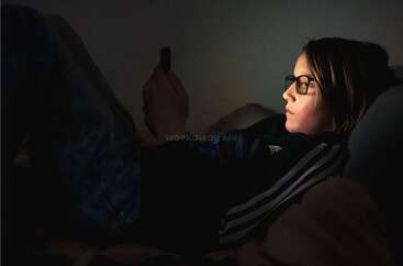Ein junger Mensch mit Brille liegt auf einem Bett in einem dunklen Raum, beleuchtet vom Licht eines Smartphones, das er in der Hand hält und auf das er schaut.