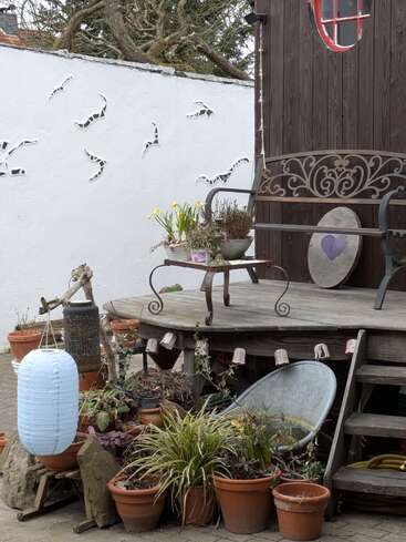 Eine gemütliche Gartenszene mit Topfpflanzen, einer dekorativen Bank, einer hängenden Laterne, Mosaikvögeln auf einer weißen Wand und rustikalen Holzelementen schafft eine charmante Atmosphäre.