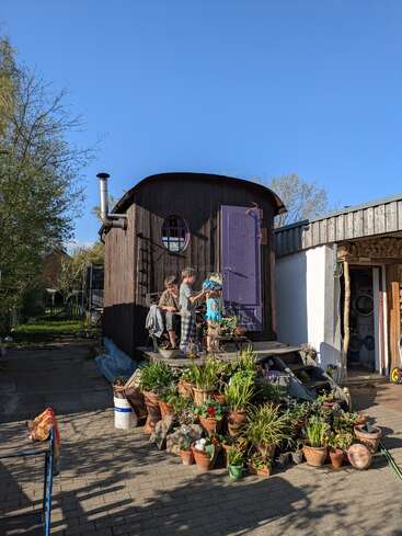 Ein kleines Holzhaus mit einer lila Tür steht im Sonnenlicht. Drei Kinder stehen auf den Stufen, umgeben von vielen Topfpflanzen und Grünzeug.
