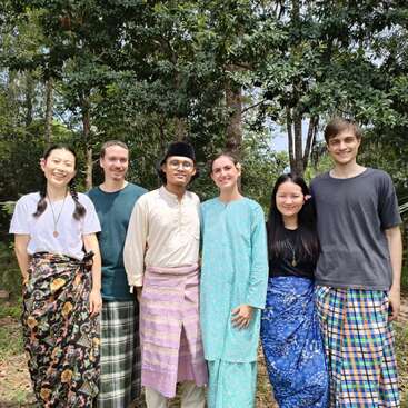 Six friends stand together outdoors, smiling happily. They wear traditional and modern attire, with sarongs, floral prints, and colorful outfits, surrounded by lush green trees and nature.