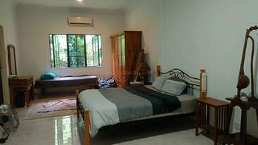 This spacious, well-lit bedroom features a double bed, single bed, wooden wardrobe, vanity table, fan, window with greenery outside, and minimalistic decor throughout.