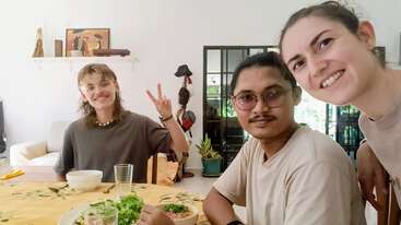 Three friends are sitting around a dining table enjoying a meal together. They are smiling, with one flashing a peace sign. The atmosphere feels warm and friendly.