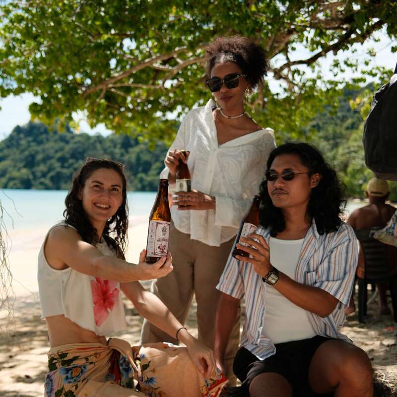 Three friends enjoy a sunny day at the beach, holding beer bottles, smiling, and relaxing under the shade of a tree with scenic water in the background.