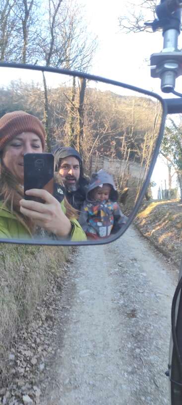 A woman, man, and baby are seen in a motorcycle mirror selfie, walking on a dirt path through a wooded, rural area on a bright day.