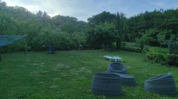 This image shows a lush green backyard with modern outdoor chairs, two white loungers, trees, a clothesline, and a swing, all under a cloudy sky.