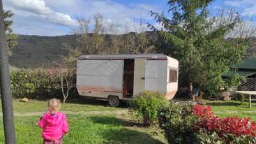 A young child in a bright pink jacket stands in a grassy yard, facing an old caravan. Trees, hills, and scattered bushes surround the peaceful scene.
