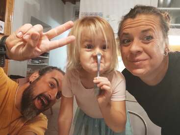 A cheerful family selfie: a man flashes a peace sign, a young girl balances a spoon on her nose, and a woman smiles warmly beside them.