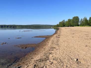 Ein ruhiges Seeufer mit glattem Wasser, Sandstrand und vereinzelten Abfällen. Grüne Bäume säumen den Horizont unter einem strahlend blauen Himmel und schaffen eine friedliche, natürliche Landschaft.