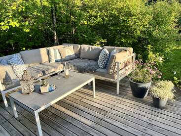 Eine gemütliche Terrasse mit einem beigen Ecksofa, gemusterten Kissen, weichen Überwürfen, einem hellen Holztisch, Getränken, einer Laterne, Topfpflanzen und einer üppig grünen Umgebung.