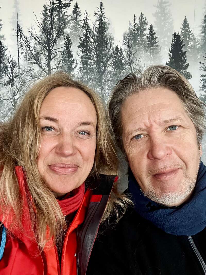 Eine lächelnde Frau und ein Mann machen gemeinsam ein Selfie aus nächster Nähe. Beide tragen Outdoor-Jacken und Schals. Hinter ihnen ist eine Kulisse aus verschneiten Bäumen und Wald zu sehen.