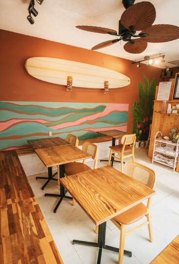 This cozy cafe features wooden tables and chairs, a surfboard wall decoration, leafy plants, and a colorful wave mural, creating a tropical, beach-inspired atmosphere.