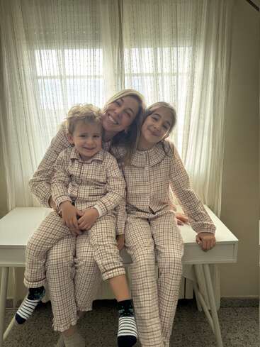 Una mujer y dos niños, todos con pijamas a juego de cuadros, están sentados juntos sonriendo en una mesa blanca delante de unas cortinas transparentes, irradiando calidez y felicidad.