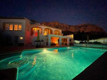 The image depicts a large white house with a pool and a mountainous backdrop at night, exuding a serene and luxurious ambiance.