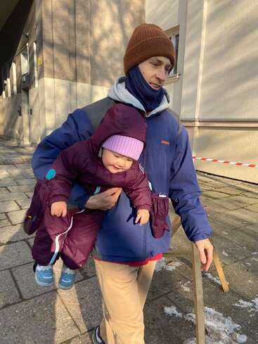 Un hombre vestido con ropa de invierno lleva en brazos a un niño pequeño, ataviado con un traje de nieve morado y un gorro rosa, en el exterior en un día soleado, pasando junto a edificios y montones de nieve.