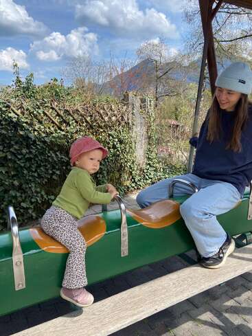 Un niño y un adulto están sentados en un balancín verde al aire libre. El niño lleva un gorro rosa; ambos parecen felices en un día soleado y parcialmente nublado.