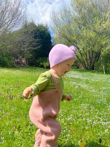 Un niño pequeño muy alegre, con un gorro rosa y un mono, explora una pradera de un verde intenso, rodeado de flores silvestres en flor y árboles, bajo un cielo luminoso y parcialmente nublado.