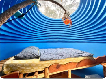 Ein gemütliches Zimmer mit hellblauen, geschwungenen Wänden, einem Bett mit Holzrahmen, einzigartiger Bettwäsche, einem runden Oberlicht und einer Hängelampe für sanftes Licht.