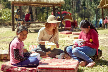 Tres personas están sentadas al aire libre sobre cojines estampados, bordando juntas. El entorno es soleado y relajado, con cabañas de paja al fondo y otras cerca, lo que crea un ambiente comunitario.