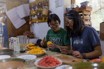 Dos mujeres preparan alegremente la comida juntas en una acogedora cocina, cortando mangos y otros ingredientes. El ambiente es cálido, agradable y está lleno de utensilios de cocina.