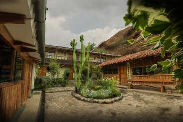 Esta imagen muestra un patio rústico con suelo de piedra, un jardín central de cactus, bancos de madera, plantas y edificios tradicionales, rodeado de exuberante vegetación y una ladera.
