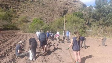 Un grupo de personas trabaja conjuntamente en un campo, cavando y plantando. El entorno es rural, rodeado de colinas, árboles y un cielo azul claro.