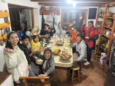 Un alegre grupo de niños y adultos se reúne alrededor de una mesa de cocina, disfrutando de la comida y la bebida, sonriendo y saludando alegremente en un ambiente acogedor y rústico.