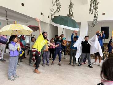 Un animado grupo de niños y un adulto actúan en un espacio interior. Sostienen coloridos paraguas y accesorios, sonríen y posan con entusiasmo, creando un ambiente alegre y festivo.