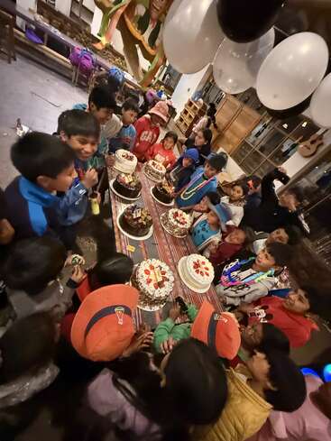 Un grupo de niños se reúne emocionado alrededor de una mesa llena de pasteles. Las decoraciones coloridas y los globos sugieren que se trata de una animada celebración de cumpleaños en un ambiente festivo.