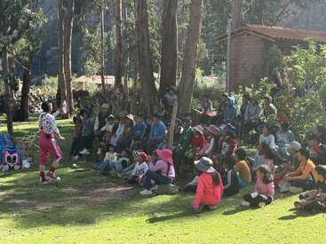 Un payaso entretiene a un numeroso grupo de niños sentados en el césped al aire libre, rodeados de árboles, con la luz del sol filtrándose a través de las hojas, creando un ambiente alegre y animado.