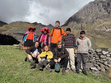 Un grupo de ocho personas, algunas con ropa tradicional andina, posan al aire libre frente a un terreno montañoso bajo un cielo nublado, todos sonrientes y relajados.