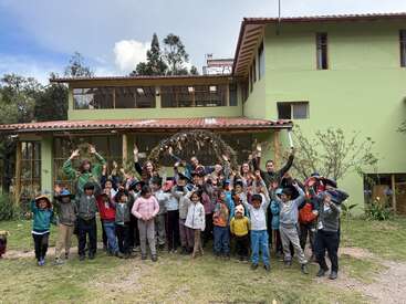 Un grupo de niños y adultos sonrientes posan juntos frente a un gran edificio verde, levantando las manos con alegría, creando un ambiente animado y alegre.