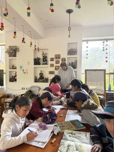 Un grupo de niños está sentado alrededor de una mesa, concentrado en escribir y dibujar, mientras un adulto les ayuda en un aula decorada y llena de color.