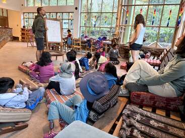 Un grupo de niños está sentado sobre cojines en una sala amplia y luminosa, escuchando a un profesor que se encuentra frente a una pizarra blanca, mientras otro adulto observa. Detrás hay grandes ventanales.