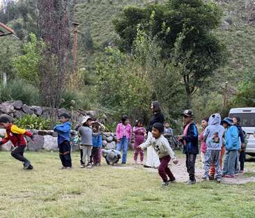 Un grupo de niños juega al aire libre en un prado, algunos corren y otros observan, rodeados de árboles y montañas, creando una escena alegre y animada.