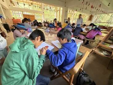Un aula llena de niños que realizan actividades en grupo, dibujan y escriben, bajo la supervisión de los profesores. La luz natural entra a raudales por los grandes ventanales, creando un ambiente cálido y animado.