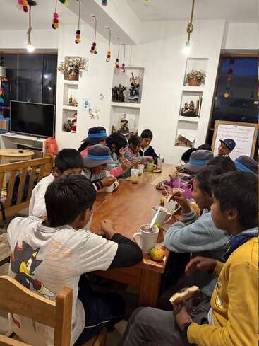 Un grupo de niños está sentado alrededor de una mesa de madera, disfrutando de aperitivos y bebidas. La sala está decorada con pompones de colores y adornos típicos, lo que crea un ambiente acogedor.