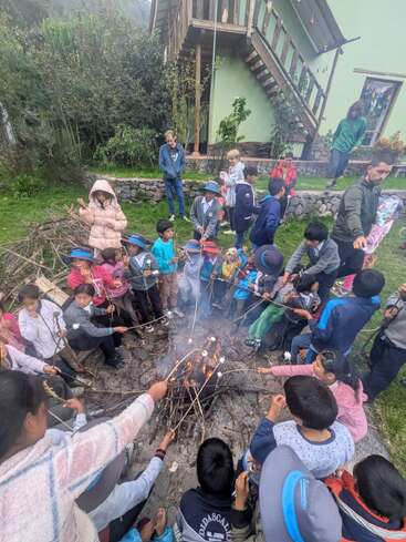 Un numeroso grupo de niños y algunos adultos se reúnen alrededor de una hoguera en un jardín, asando malvaviscos en palitos y disfrutando juntos de una actividad al aire libre.