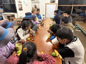 Un grupo de niños y adultos está sentado alrededor de una mesa de madera, disfrutando de aperitivos y bebidas, charlando y relacionándose en un ambiente acogedor en el interior, con una pizarra blanca.
