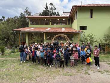 Un numeroso grupo de adultos y niños posa juntos frente a un edificio verde con techo rojo, rodeado de árboles y montañas, mientras disfrutan de una reunión comunitaria.