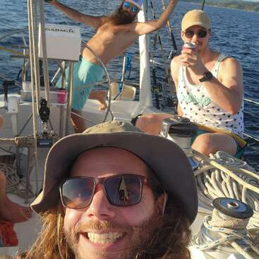 Três amigos estão sorrindo e se divertindo em um veleiro, aproveitando o clima ensolarado, as bebidas e as águas abertas, criando uma atmosfera alegre e descontraída.