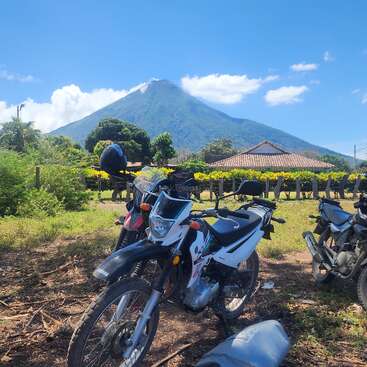 Três motocicletas estão estacionadas em uma área gramada, com um cenário de um vulcão, vegetação exuberante, uma casa com telhado de telha e céu azul.