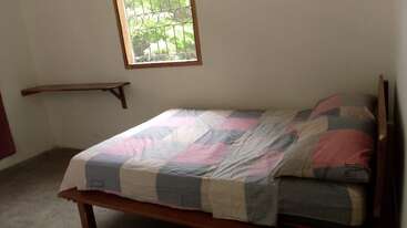 Esta imagem mostra um quarto simples com uma cama de casal, roupa de cama xadrez, uma moldura de madeira, uma pequena janela com grades e uma prateleira de canto na parede.
