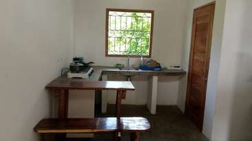 Esta imagem mostra uma cozinha simples e pequena com uma mesa e um banco de madeira, um fogão, panelas, uma pia sob uma janela e uma porta de madeira.
